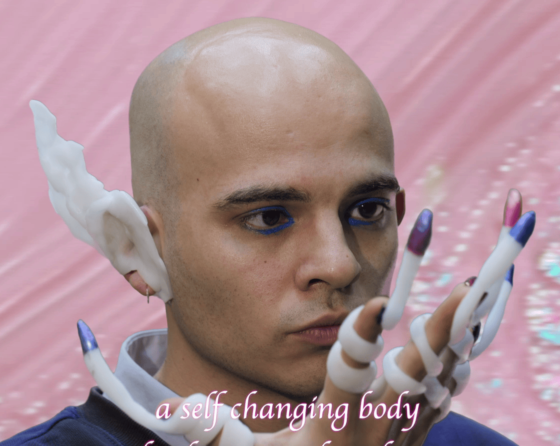 Filmstill of Milan Gress with a shaved head and blue eyeliner gazes  at their hand, which is adorned with sculptural, elongated, white and colorful finger extensions. They also wear a large, white, sculpted ear accessory. The background is a soft, pastel pink with subtle sparkles, and part of the phrase 'a self changing body' is visible at the bottom in pink text.